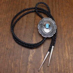 May include: A silver bolo tie with a turquoise stone set in a silver oval pendant. The tie is on a black braided cord.