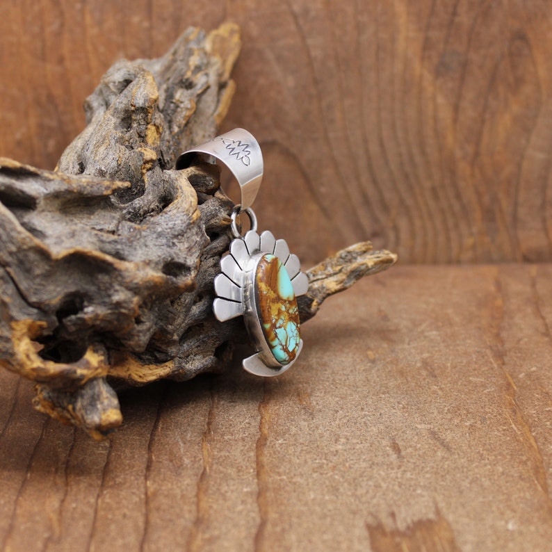 May include: A silver pendant with a turquoise and brown stone set in a silver design. The pendant has a silver bail with the word "CWM" engraved on it.