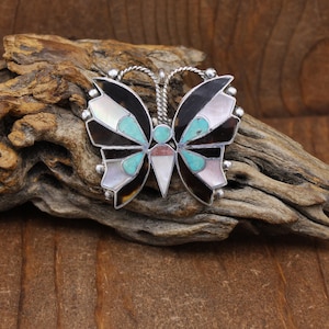 May include: A silver butterfly pendant with inlaid turquoise, mother-of-pearl, and black onyx. The butterfly design features a detailed wing pattern and a twisted silver bail for a necklace chain. The pendant is set against a natural wood background.