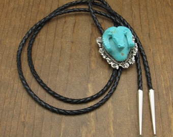 May include: A turquoise blue bolo tie featuring a carved ram's head with silver accents. The tie is made with black braided leather cord and silver tips.