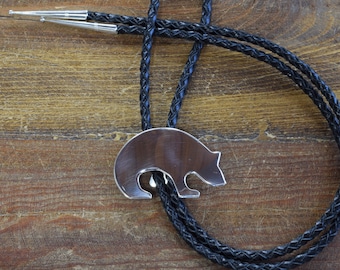 May include: A silver bolo tie featuring a bear pendant with a dark brown center. The bear pendant is connected to a black braided cord with silver tips. The background is a wooden surface.