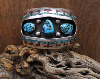 May include: A silver cuff bracelet with turquoise and red-brown inlay. The bracelet features three large turquoise stones and a geometric design around the edges. The bracelet is displayed on a piece of driftwood.