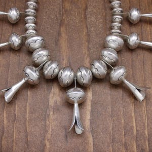 May include: A silver necklace with a central pendant and multiple smaller pendants. The necklace is made of round beads with a textured surface.