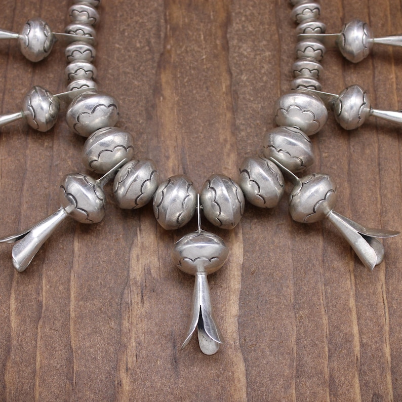 May include: A silver necklace with a central pendant and multiple smaller pendants. The necklace is made of round beads with a textured surface.