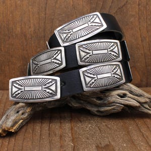 May include: A black leather belt with five rectangular silver buckles. Each buckle features a sunburst design with a central rectangular element and geometric patterns. The belt is displayed on a wooden surface.