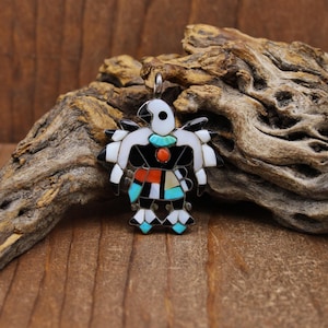 May include: A handcrafted pendant in the shape of a bird, featuring inlaid stones in black, white, turquoise, red, and tan. The bird has a silver bail for hanging. The pendant is set against a natural wood background.