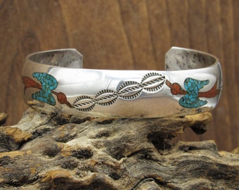 May include: A silver bracelet with turquoise and red stones set in a geometric pattern. The bracelet is decorated with a repeating design of stylized birds.