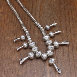 May include: A silver necklace with a beaded chain and silver pendant drops. The beads are round and have a textured surface. The pendant drops are shaped like teardrops and have a smooth surface.