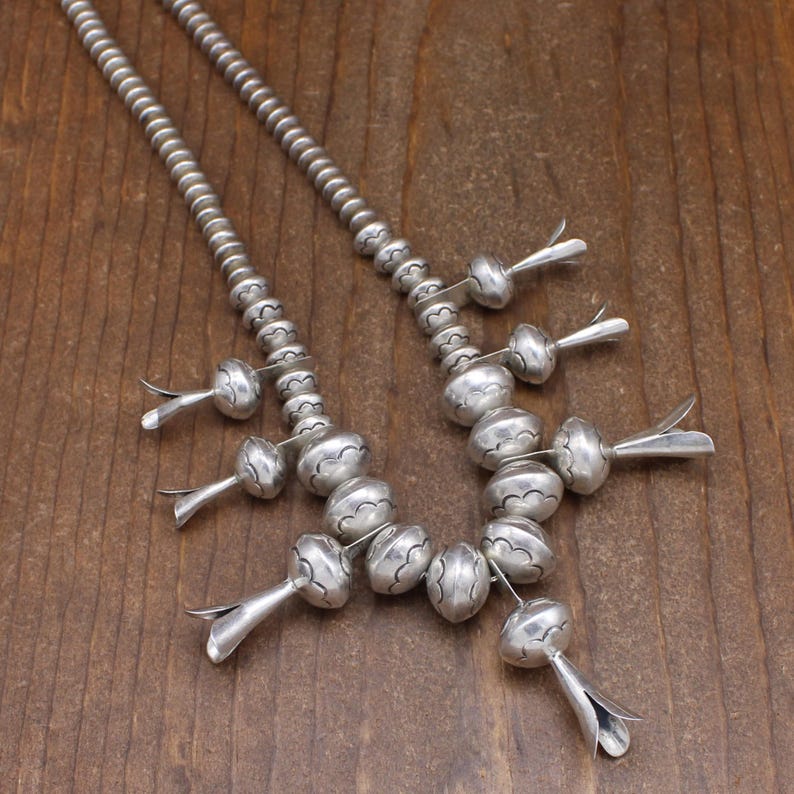 May include: A silver necklace with a beaded chain and silver pendant drops. The beads are round and have a textured surface. The pendant drops are shaped like teardrops and have a smooth surface.