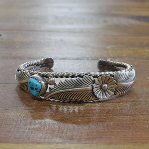 May include: Silver cuff bracelet with a turquoise stone and floral and feather designs. The bracelet has a braided rope design on the sides. The turquoise stone is a light blue color. The bracelet is on a wooden surface.