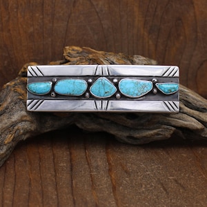 May include: A rectangular silver belt buckle featuring five turquoise stones. The stones are set in a row and are various shapes. The silver buckle has a geometric design with etched lines and is displayed on a wooden surface.