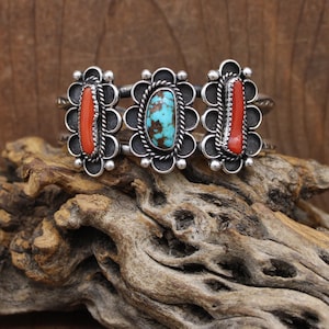 May include: A silver cuff bracelet featuring three decorative floral settings. The settings hold oval-shaped gemstones: two red coral and one turquoise. The bracelet is displayed on a piece of driftwood.