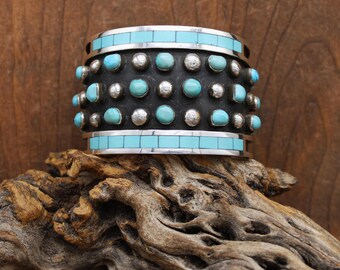 May include: A wide silver cuff bracelet with turquoise and silver accents. The bracelet features a band of turquoise stones at the top and bottom, with rows of turquoise and silver studs in the center. The bracelet is displayed on a piece of driftwood.