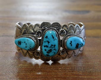 May include: A silver cuff bracelet with three turquoise stones set in the center. The bracelet has a decorative design with scalloped edges.