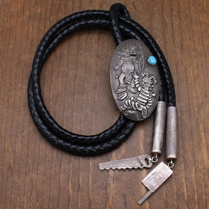 May include: A bolo tie with a black braided leather cord. The silver-toned pendant features an engraved design of two figures and a turquoise stone. The tie also includes silver-toned metal tips and decorative saw and cleaver charms.