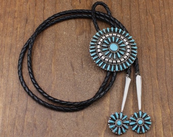 May include: A turquoise and silver bolo tie with a large circular pendant and two smaller circular accents. The pendant is made of silver and features a turquoise inlay in a sunburst pattern. The bolo tie is made of black leather cord.
