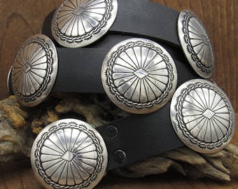 May include: A black leather belt with five silver concho belt buckles. Each buckle has a raised, diamond-shaped design in the center.
