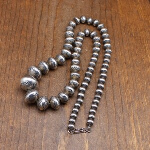 Puede incluir: Un collar de plata con una cadena de cuentas y un dise&ntilde;o de cuentas m&aacute;s grandes grabadas. El collar est&aacute; colocado sobre una superficie de madera.