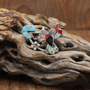 May include: A silver-toned hummingbird brooch with inlaid stones. The bird features red, black, and iridescent accents, with turquoise flower and branch details. The brooch is set against a natural wood background.