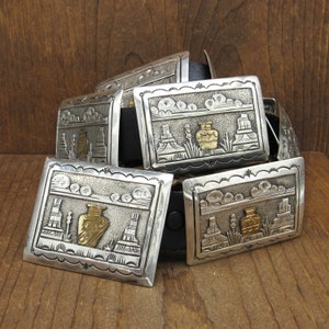 May include: A silver belt buckle with a southwestern design featuring a gold-colored vase and stylized buildings. The buckle is part of a leather belt.