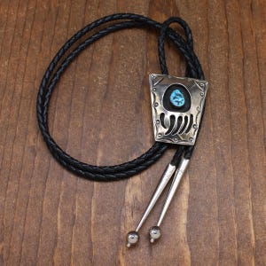 May include: A silver bolo tie with a turquoise stone set in a bear paw design. The bolo tie has a black leather cord.