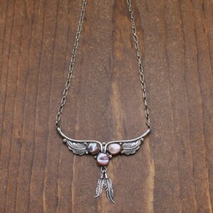 May include: A silver necklace with a delicate chain and a pendant featuring three pink abalone shell stones and two silver feathers. The pendant is shaped like a bird with outstretched wings.