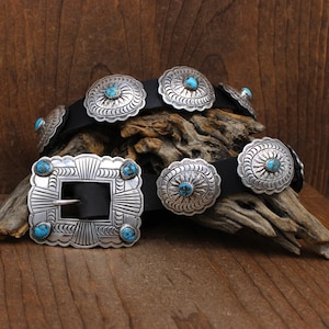 May include: A black leather belt with silver and turquoise concho belt buckles. The belt is folded and displayed on a piece of driftwood.