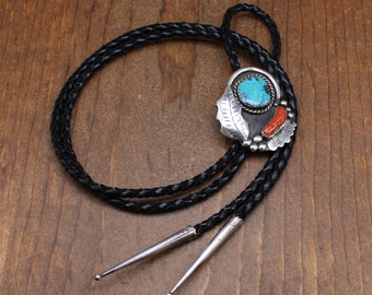 May include: A silver bolo tie featuring a turquoise and coral stone design. The bolo tie has a black leather cord and silver tips.