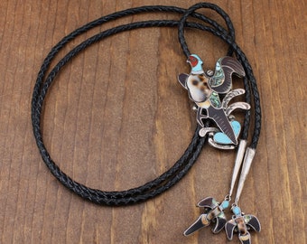 May include: A black leather bolo tie with a decorative bird pendant. The bird design incorporates a speckled body, turquoise details, and a red head. The tie includes two matching bird-shaped tips.
