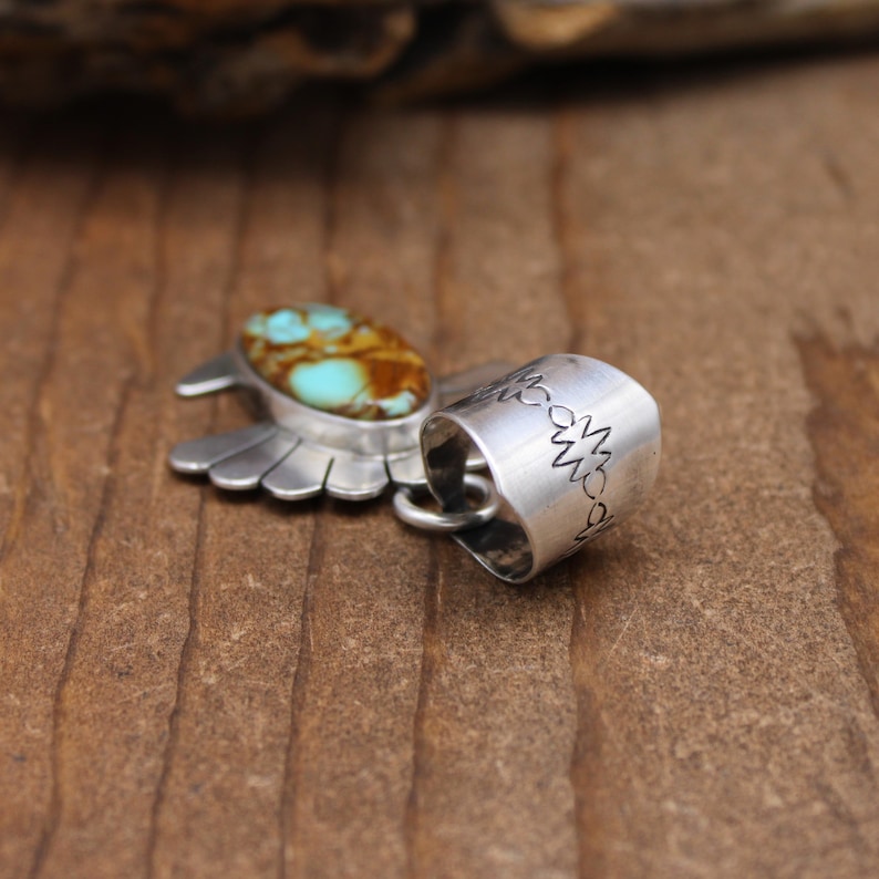 May include: Silver pendant featuring a turquoise stone set in a stylized wing design. The pendant has a decorative bail with a repeating pattern.