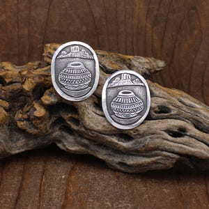 May include: Two oval-shaped silver earrings. Each earring features a detailed design of a pot, mountains, and water. The earrings are set against a natural wood background, showcasing their intricate craftsmanship and cultural theme.