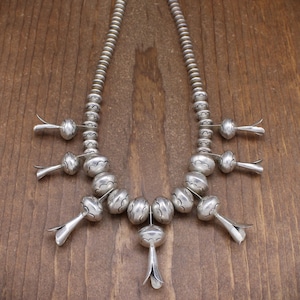 May include: A silver necklace with a repeating pattern of round beads and flower-shaped pendants. The necklace is made of sterling silver and has a traditional Native American design.