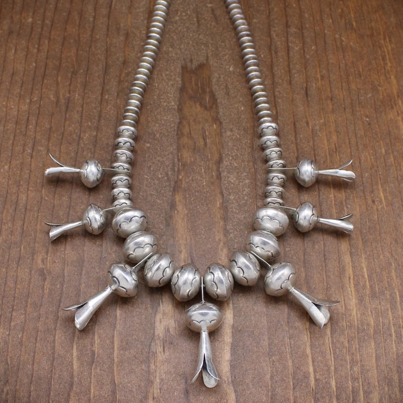 May include: A silver necklace with a repeating pattern of round beads and flower-shaped pendants. The necklace is made of sterling silver and has a traditional Native American design.