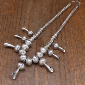 May include: A silver necklace with a cluster of large, round beads and smaller, teardrop-shaped beads. The beads are decorated with a repeating pattern of small circles.