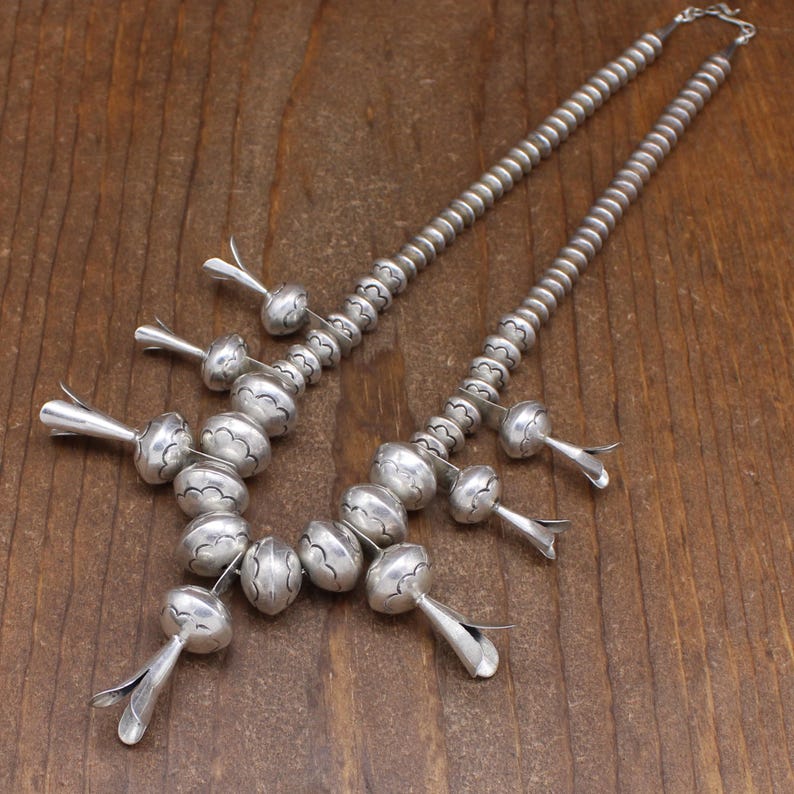 May include: A silver necklace with a cluster of large, round beads and smaller, teardrop-shaped beads. The beads are decorated with a repeating pattern of small circles.