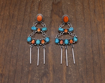 Peut inclure: Une paire de boucles d'oreilles chandelier en argent avec des pierres turquoise et orange en forme d'ovale et de goutte. Les boucles présentent un design à étages avec des accents en fil d'argent et trois barres argentées pendantes.
