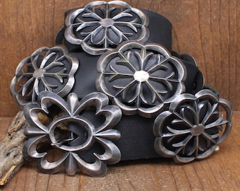 May include: A black leather belt with five silver concho belt buckles. The buckles are all different designs, but they all have a floral motif. The belt is lying on a wooden surface.
