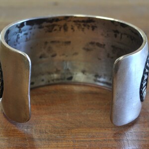 K&ouml;nnte beinhalten: Ein silbernes Armreif mit geometrischem Muster. Das Armband ist an einer Seite offen und hat eine glatte, polierte Oberfl&auml;che.