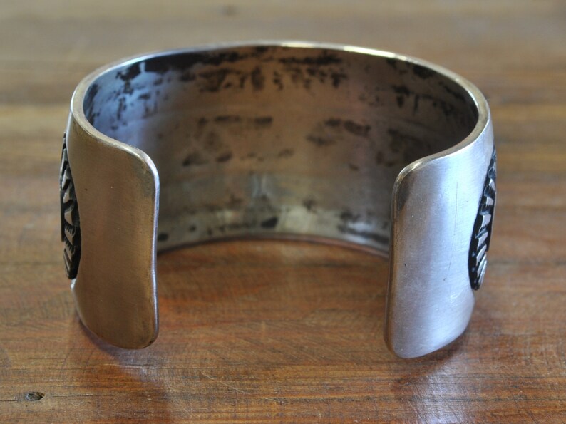 K&ouml;nnte beinhalten: Ein silbernes Armreif mit geometrischem Muster. Das Armband ist an einer Seite offen und hat eine glatte, polierte Oberfl&auml;che.