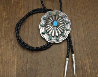 May include: A silver bolo tie with a turquoise stone in the center and a braided black leather cord.