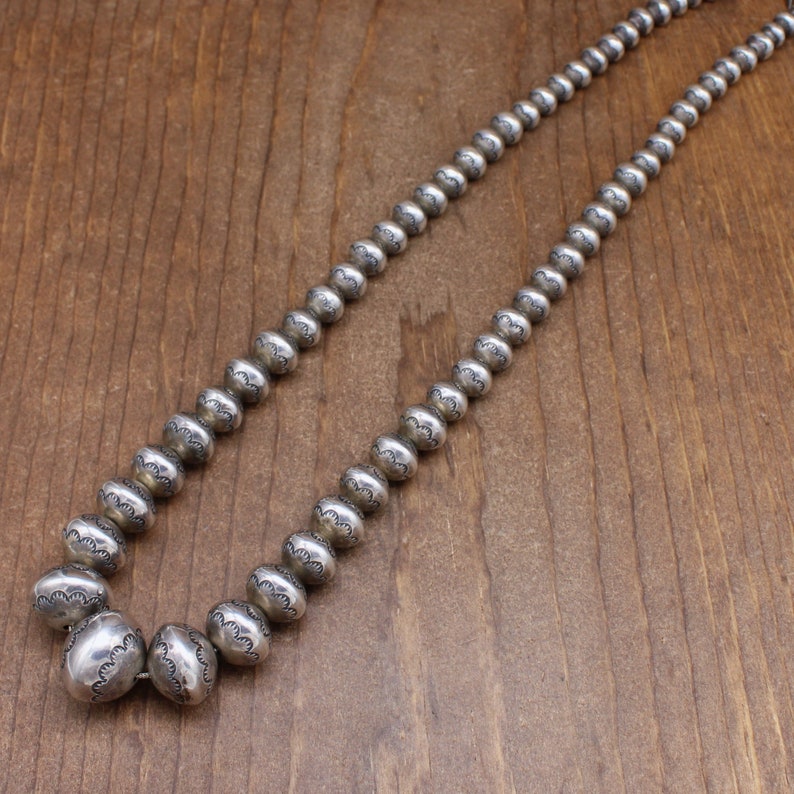 Puede incluir: Un collar de plata con cuentas redondas, cada cuenta tiene un patr&oacute;n decorativo. El collar est&aacute; sobre una superficie de madera.