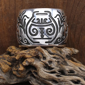 May include: A wide, silver cuff bracelet with a detailed, etched design. The design features a central, stylized pot shape with swirling patterns and linear elements. The bracelet is displayed on a piece of driftwood.