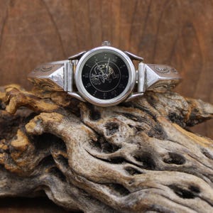 May include: A silver bangle watch with a black face and Roman numeral hour markers. The watch face features a stylized Native American dancer design.