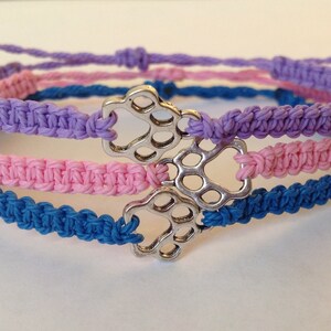 May include: Macrame bracelets in purple, pink, and blue, each featuring a silver paw print charm. The bracelets are adjustable and appear to be handmade, perfect for pet lovers.