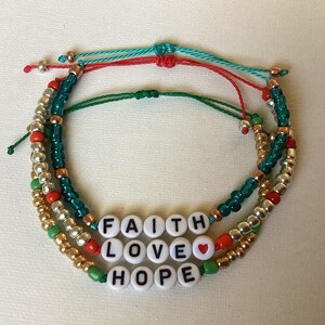 May include: Three layered bracelets with green, red, and gold beads. The bracelets spell out "FAITH LOVE HOPE" with a small red heart bead between "LOVE" and "HOPE".