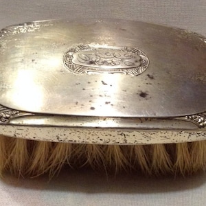 May include: A silver-plated hairbrush with an ornate design and a monogrammed oval on the top. The brush has a worn, vintage look.