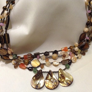 Peut inclure: Collier multi-rangs avec des perles brunes, beiges et orange, et un pendentif en forme de larme en coquille irisée.