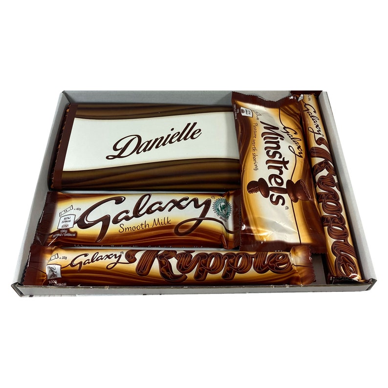 May include: A gift box filled with chocolate bars. The box is white and has a brown lid. The chocolate bars are wrapped in brown and gold foil. The chocolate bars are Galaxy Smooth Milk, Galaxy Ripple, and Cadbury Minstrels. The box is personalized with the name "Danielle".
