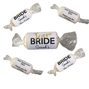 May include: Four white favor boxes wrapped in clear cellophane with gold and black text. The boxes have different designs, including "Bride Sarah's Hen Party", "Team Bride Sarah's", and "24th September 2022 Thank you for coming".
