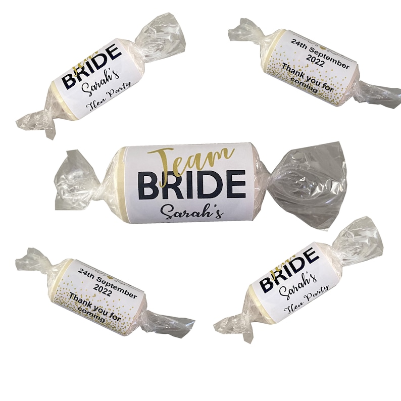 May include: Four white favor boxes wrapped in clear cellophane with gold and black text. The boxes have different designs, including "Bride Sarah's Hen Party", "Team Bride Sarah's", and "24th September 2022 Thank you for coming".